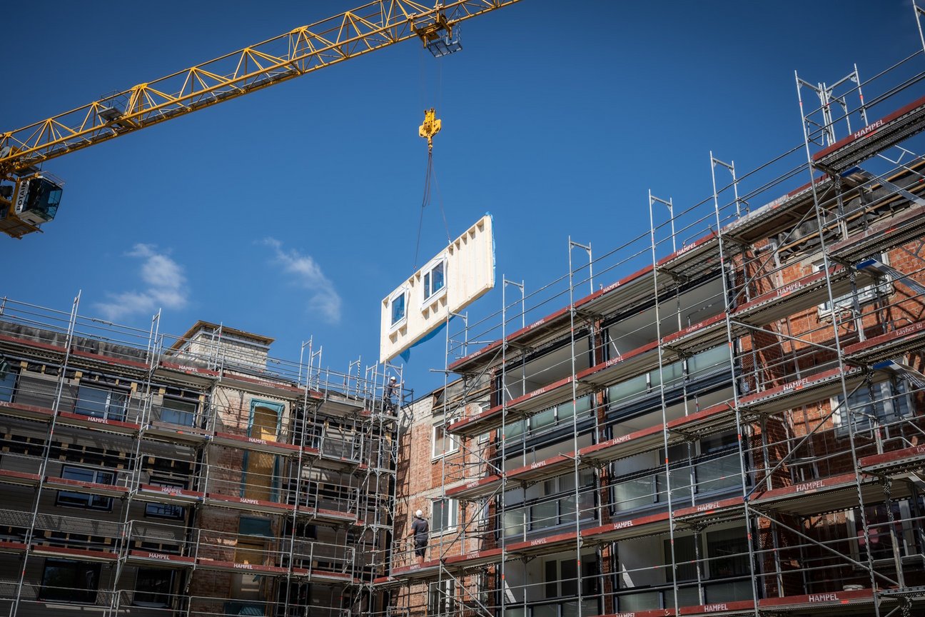 Gebäude mit Baugerüst während Sanieurng, vorgefertigtes Fassadenelement mit Fenstern schwebt davor an Kran.
