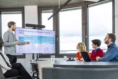Vier Personen sitzen am Tisch in einem Besprechungsraum, eine Person steht und erklärt etwas am Bildschirm, im Hintergrund befinden sich Fenster.