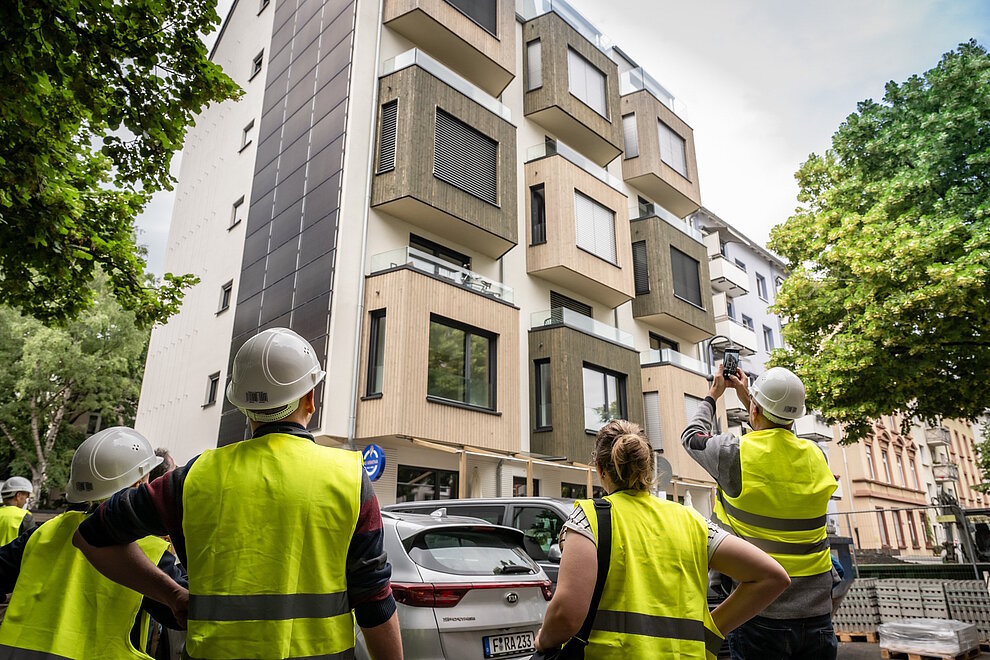 Personen mit Bauhelmen und Sicherheitswesten schauen fertig saniertes Mehrfamilienhaus an.
