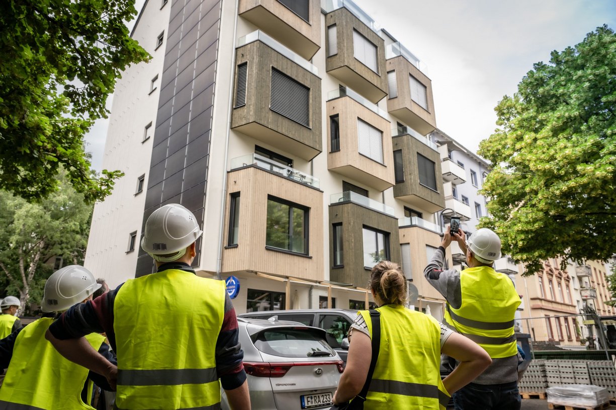 Personen mit Bauhelmen und Sicherheitswesten schauen fertig saniertes Mehrfamilienhaus an.