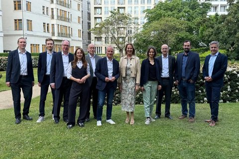 Elf Personen in Business-Kleidung stehen nebeneinander auf dem Rasen für ein Gruppenbild, im Hintergrund mehrgeschossige Gebäude