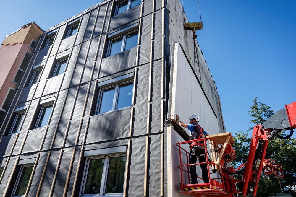 Bild zeigt Fassade eines Gebäudes und an der Seite eine Fassadenmontage mit Hebebühne und vorgefertigtem Fassadenelement