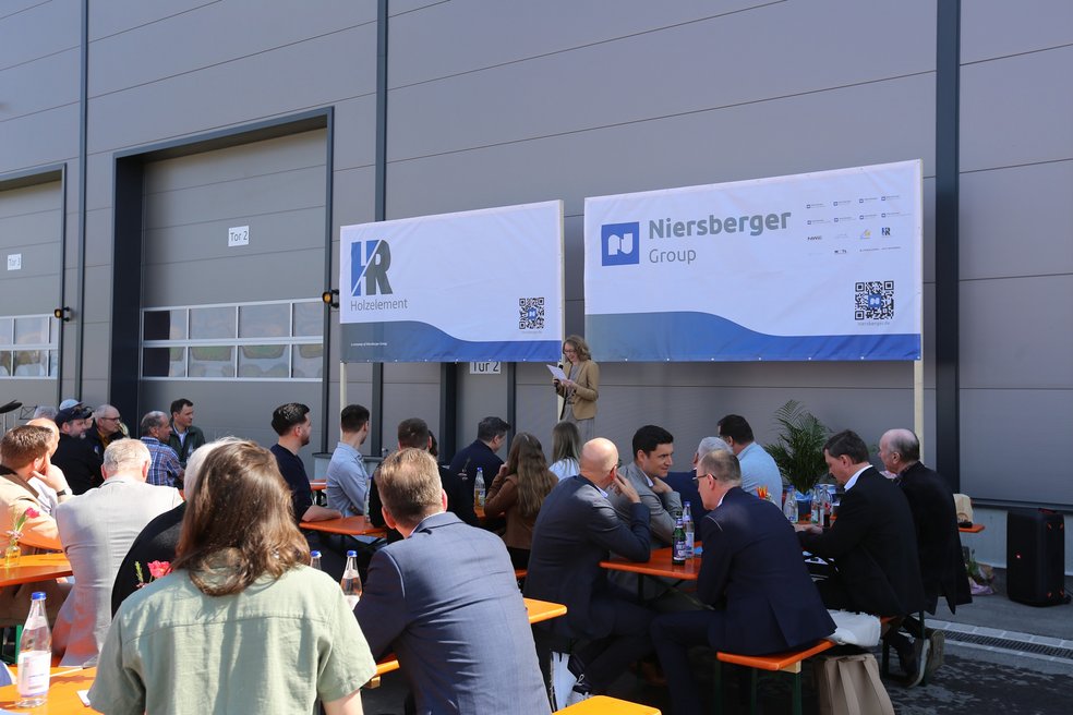 Personen sitzen an Biergartentischen und schauen zur Rednerin, dahinter die Fassade einer Produktionshalle.