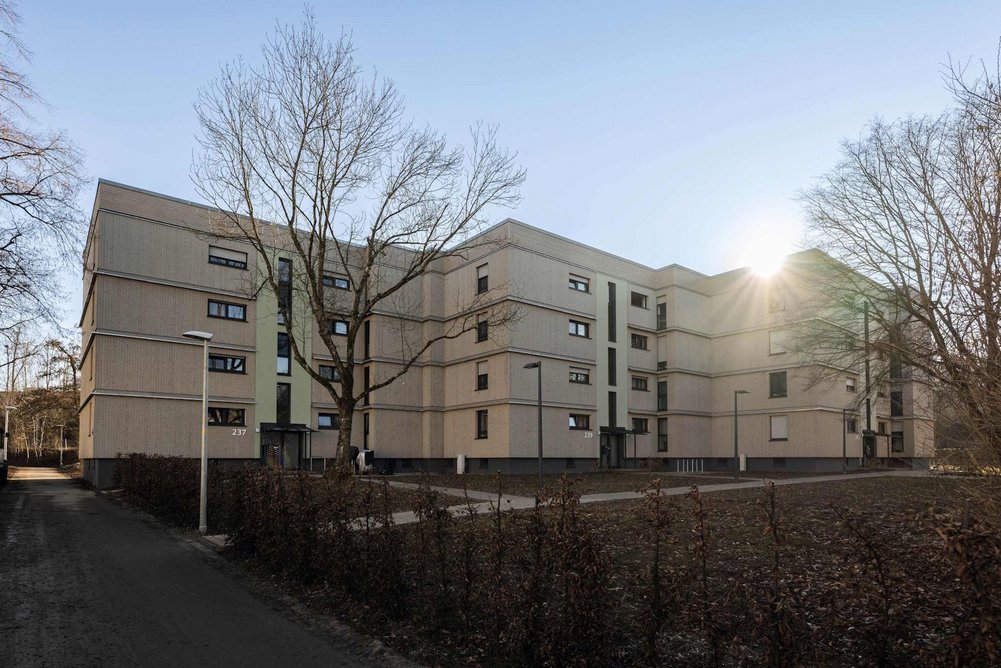 Wohngebäude viergeschossig mit Flachdach, im Hintergrund blauer Himmel, davor Grünanlagen mit Bäumen