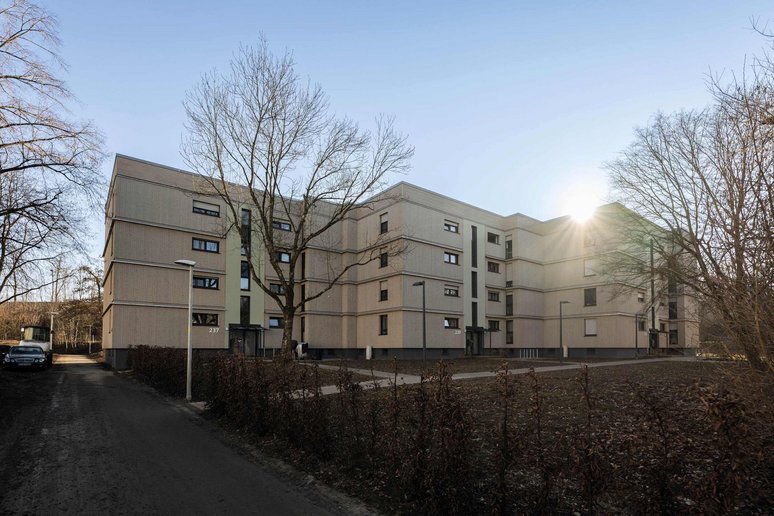 Wohngebäude viergeschossig mit Flachdach, im Hintergrund blauer Himmel, davor Grünanlagen mit Bäumen