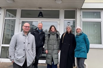 Fünf Personen in warmer Kleidung stehen nebeneinander vor einem Gebäudeeingang. 