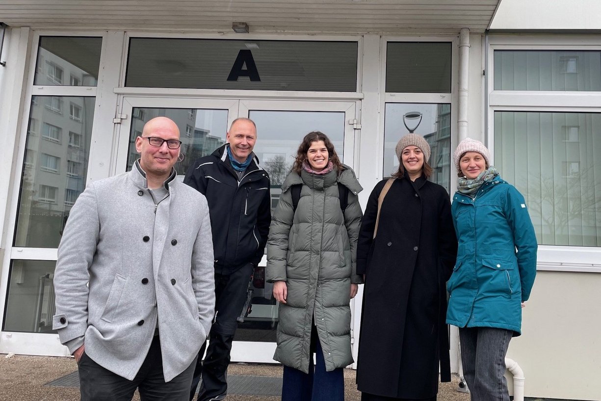 Fünf Personen in warmer Kleidung stehen nebeneinander vor einem Gebäudeeingang. 