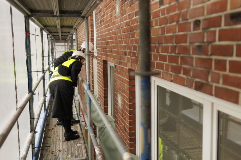 Zwei Personen stehen auf einem Baugerüst und schauen von außen in ein Fenster des Gebäudes, das saniert wird.