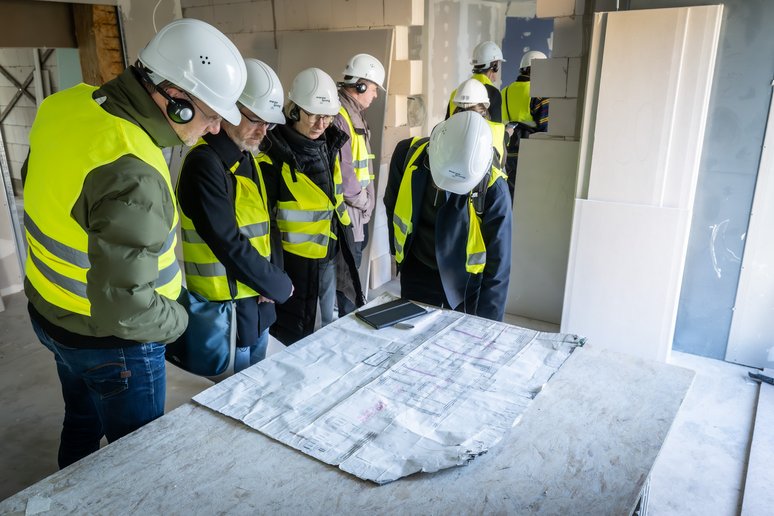 Personen mit geleben Sicherheitswesten und weißen Bauhelmen auf dem Kopf stehen in einem Gebäude und schauen auf einen Plan, der auf einem Tisch liegt.