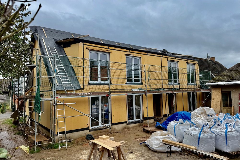In Tentzerow saniert die HALM GmbH ein Einfamilienhaus seriell mit Strohdämmung. Bild zeigt Haus mit gelber Fassade in Gerüst davor Baumaterialien