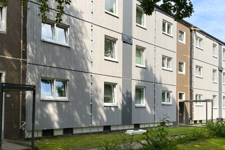 Wohngebäude mit grauer Holzfassade und Gerüst an der Seite, umgeben von einem gepflasterten Innenhof mit Grünflächen und Sträuchern.