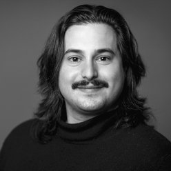 Portraitfoto in Schwarz-Weiß, Person mit dunklen, schulterlangen Haaren und Oberlippenbart