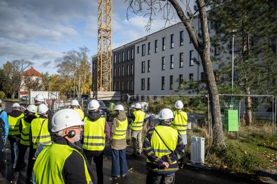 Personengruppe in geleben Sicherheitswesten und mit weißen Helmen auf dem Kopf stehen vor einer Baustelle. Im Hintergrund ein Mehrfamilienhaus während Sanierung.