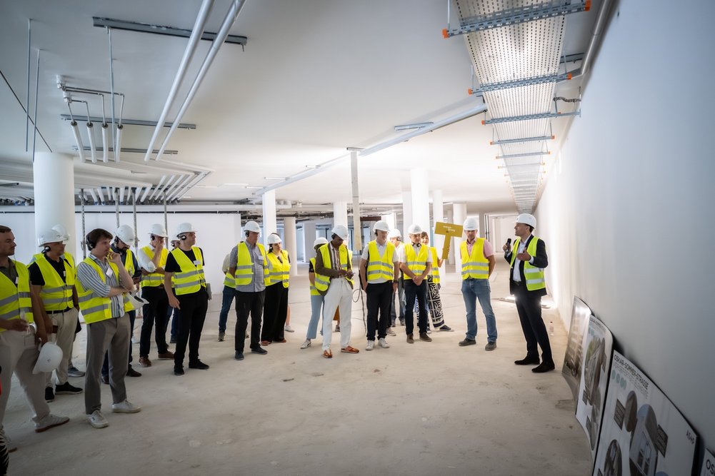 Großer Raum in einem Gebäude, dass saniert wird. Mehrere Personen mit gelben Sicherheitswesen stehen in einem Halbkreis und hören dem Architekten bei Erläuterungen zu.