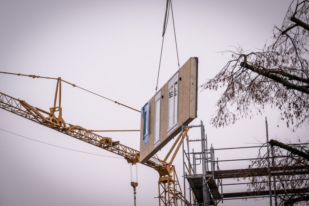 Bild zeigt: Kran mit vorgefertigtem Fassadenelement vor bewölktem Himmel