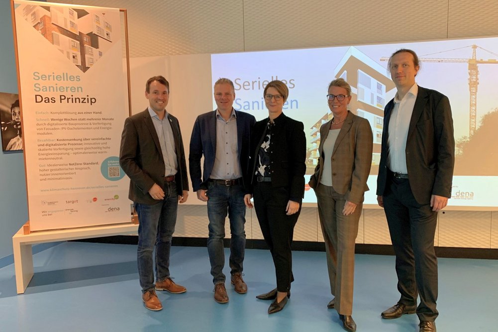 In enger Kooperation mit der Deutsche Energie-Agentur (dena) will die Klimaschutzagentur Region Hannover gemeinsam mit den Projektpartnern target, hannoverimpuls und proKlima die Region Hannover zur Vorreiterin für Serielles Sanieren in Niedersachsen machen. 5 Personen stehen vor Rollup und beleuchteter Wand mit Beamer-Präsentation