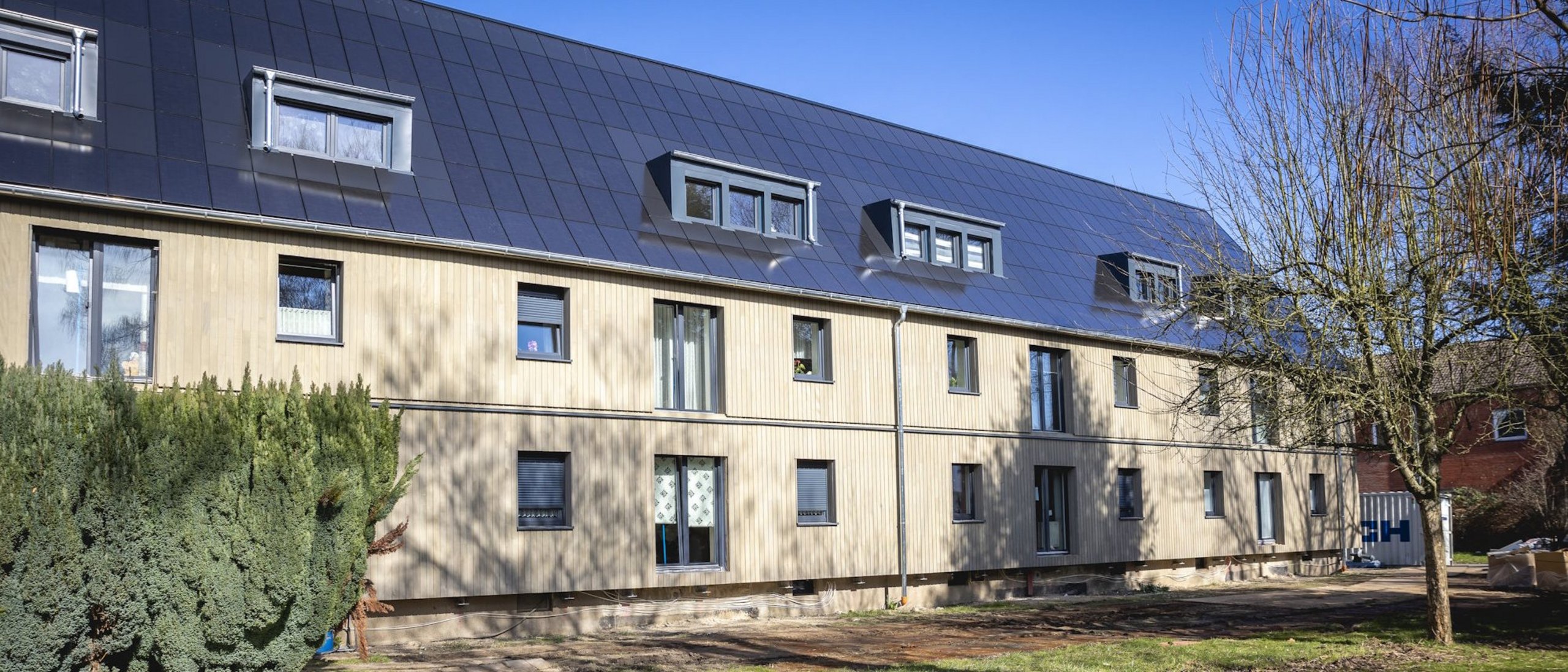 Mehrfamilienhaus zweigeschossig mit Solardach, davor Grünfläche mit Spielplatz, blauer wolkenloser Himmel
