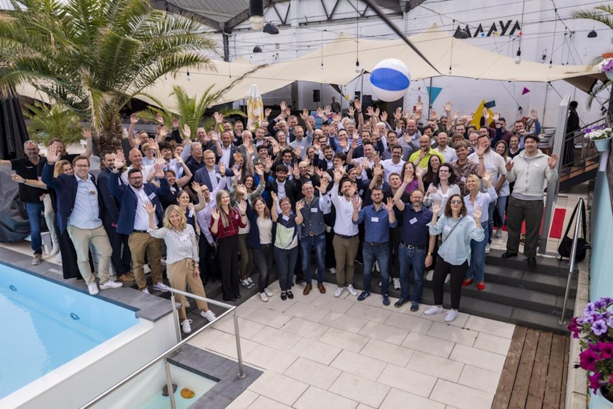 Viele Personen stehen neben einem Pool outdoor und winken.