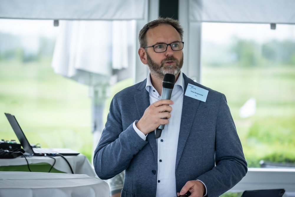 Timo Sengewald, dena-Kompetenzzentrum Serielles Sanieren Sprechende Person mit Mikrofon in der Hand, im Hintergrund Fenster