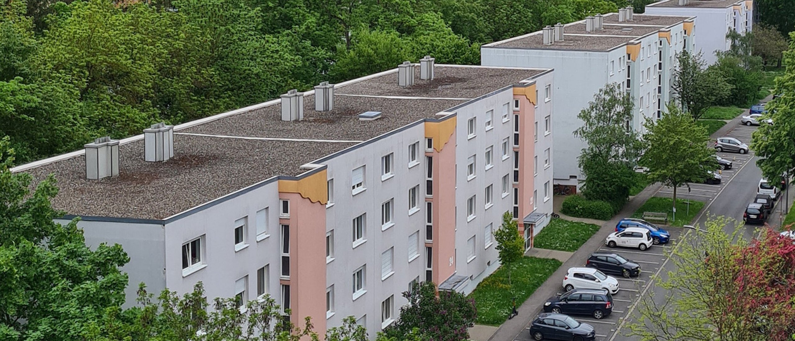 Vogelperspektive auf eine Wohnsiedlung mit mehreren weiß-grauen Mehrfamilienhäusern, begrünten Außenanlagen und einer Straße mit parkenden Autos. Im Hintergrund sind weitere Wohnhäuser und dichtes, grünes Baumwerk zu sehen.