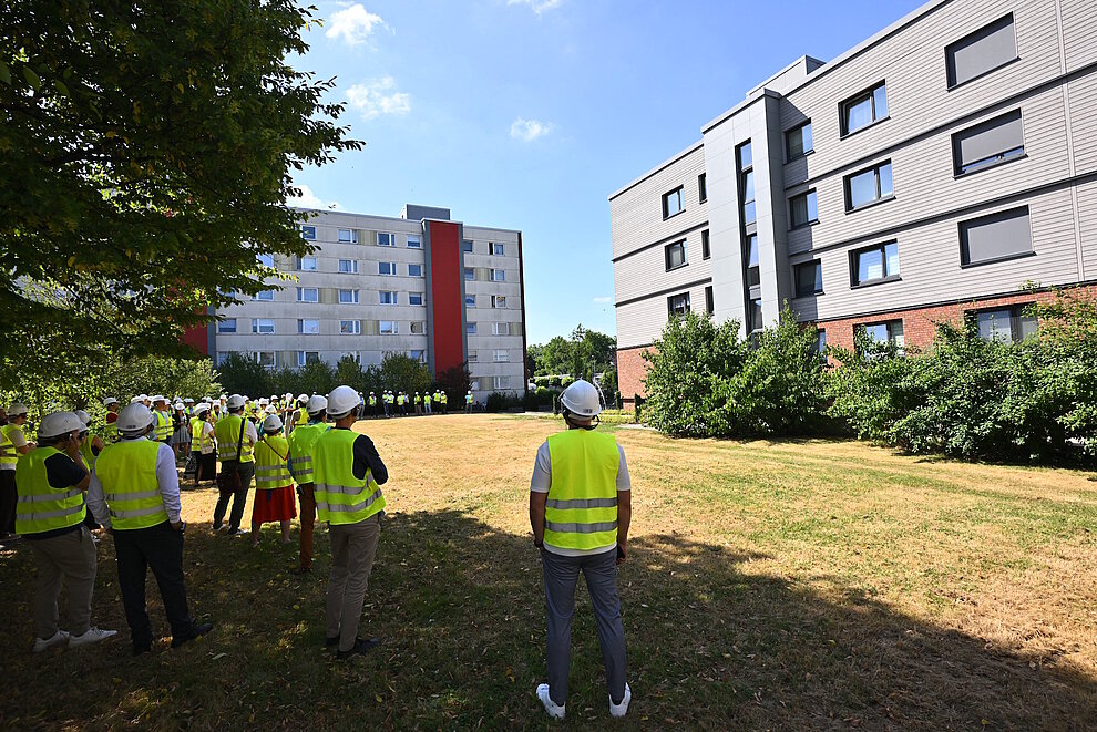 Personen mit gelben Sicherheitswesten stehen auf einer Rasenfläche vor einem sanierten Mehrfamilienhaus.