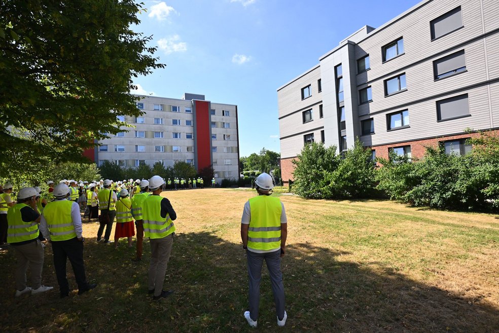Personen mit gelben Sicherheitswesten stehen auf einer Rasenfläche vor einem sanierten Mehrfamilienhaus.